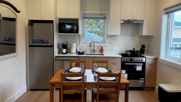 A modern kitchen with white cabinets, a built-in microwave, a coffee maker, and a stove. There is a dining table with chairs and a window above it. - New 2BR Garden Suite with AC and private patio (Burnaby)