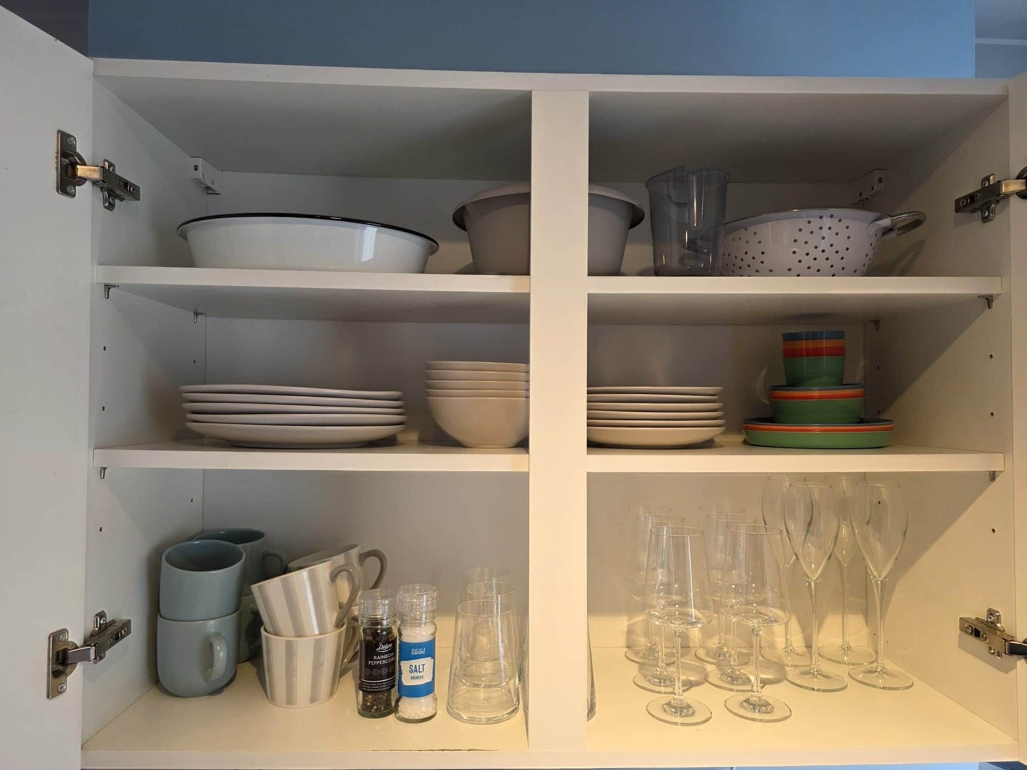 A kitchen cabinet with shelves containing plates, bowls, mugs, and wine glasses.