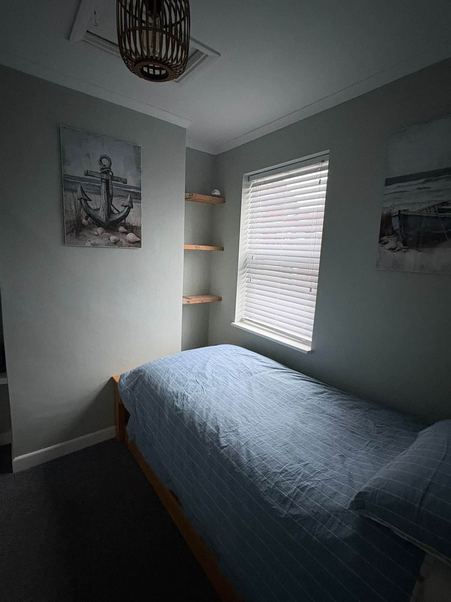 A bedroom with a single bed, a window with blinds, and a wall-mounted shelf.