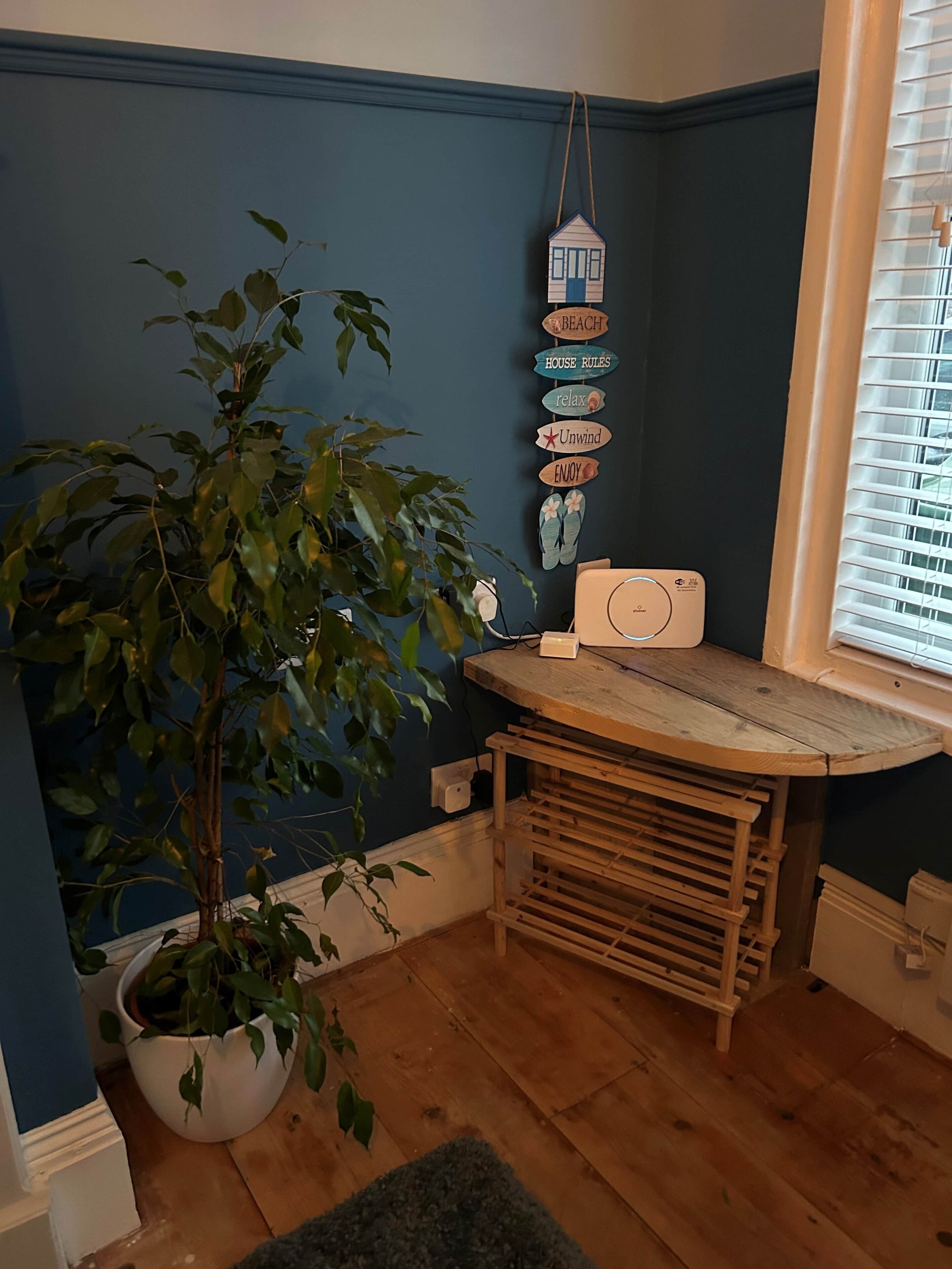A corner room with a potted plant, a wooden bench, a small table, and a wall-mounted sign with various phrases.