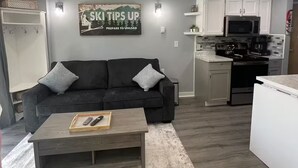 Ein modernes Wohnzimmer mit einem dunklen Sofa, einem Couchtisch und einer Küchenzeile mit weißen Schränken und einer Mikrowelle.