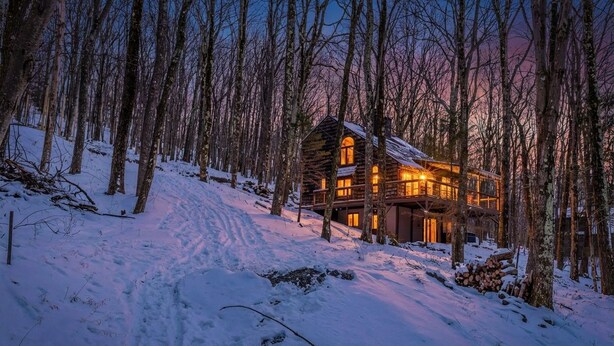 Een vakantiehuis in blokhutstijl met een ruime veranda en meerdere ramen, omgeven door een besneeuwd bos.