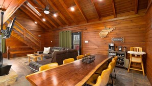 Een houten chalet van binnen, met een eettafel, stoelen, een bank, een koffiehoekje en een open haard.