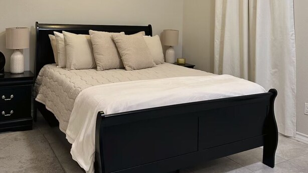 Une chambre à coucher avec un lit noir, du linge de lit blanc, deux tables de chevet avec des lampes, un ventilateur de plafond et un rideau blanc.