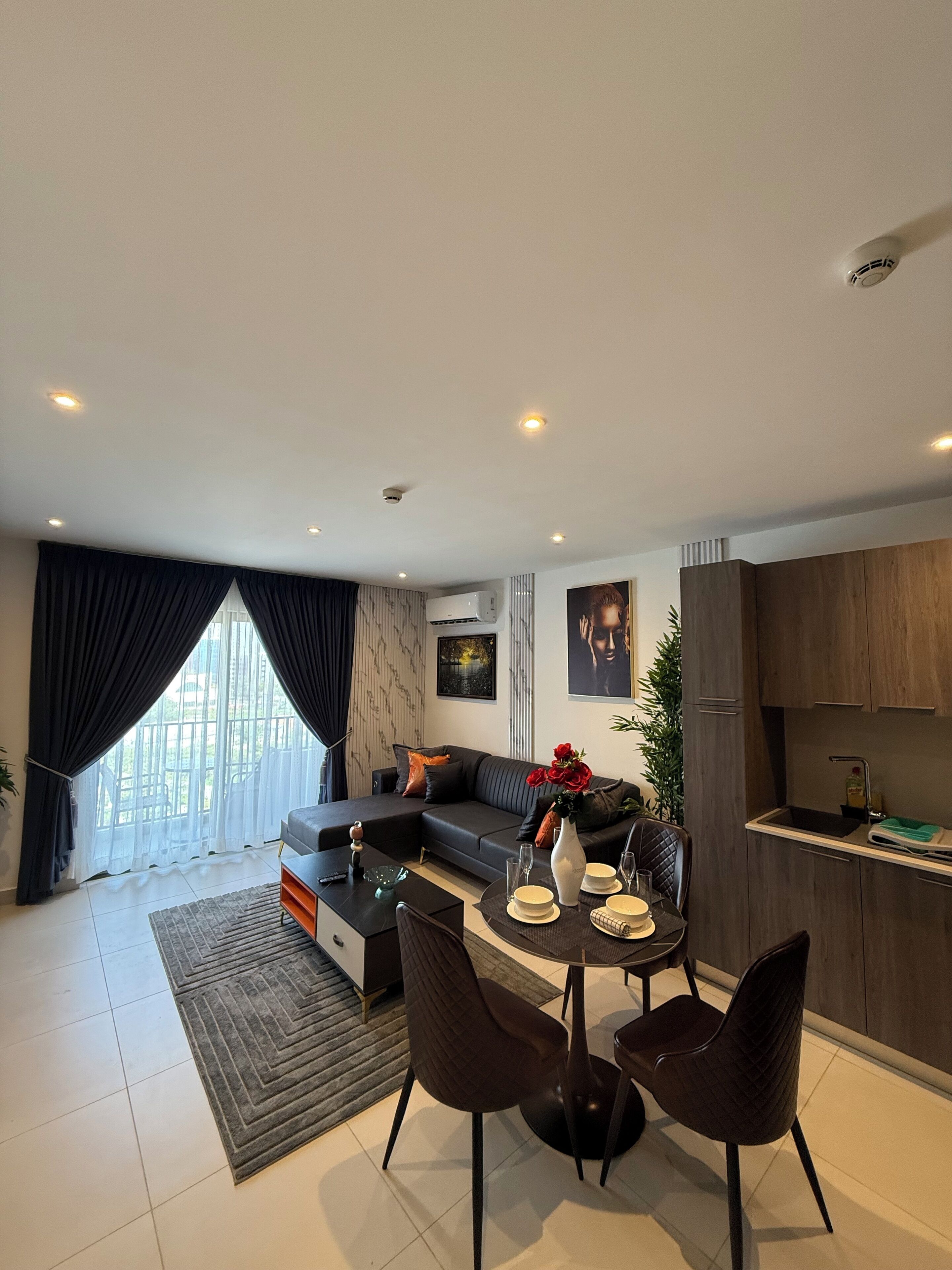 A modern living room with a sofa, dining table, and chairs, a kitchen area with wooden cabinets, and a large window with curtains.
