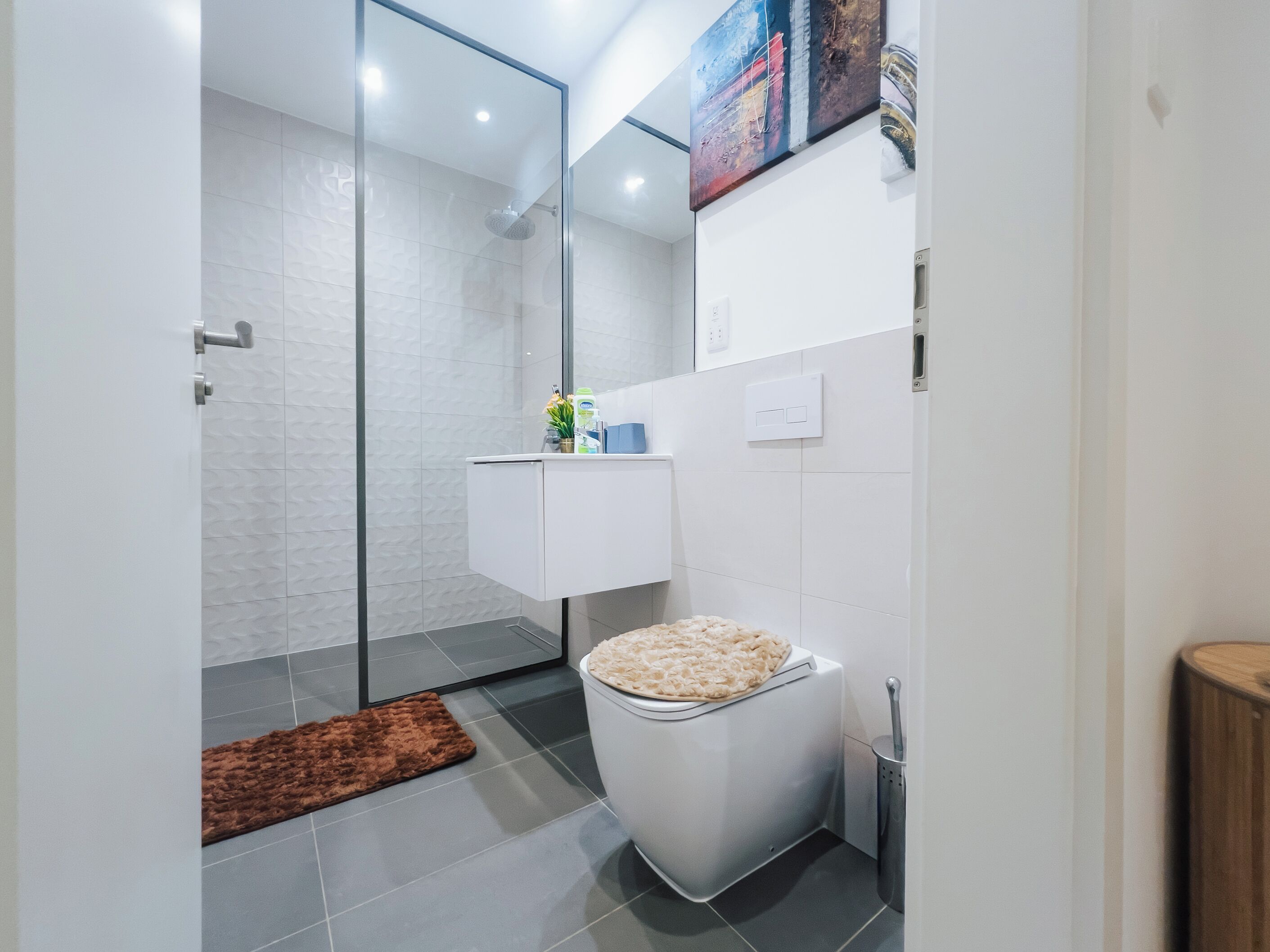 A modern bathroom with a glass shower enclosure, a white toilet, a small sink, and a brown rug on the floor.