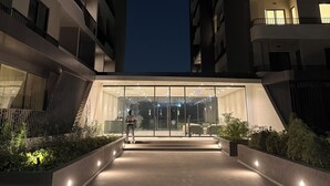 Um edifício moderno com entrada de vidro, rodeado de vegetação e iluminado por luz ambiente.