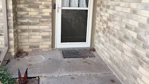 Fachada de ladrillo con puerta enmarcada en blanco, una cámara de seguridad y un pequeño jardín con una placa de metal decorativa.