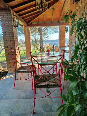 Een overdekt terras met een glazen tafel en rode metalen stoelen, met uitzicht op een tuin met bomen en een landschap.