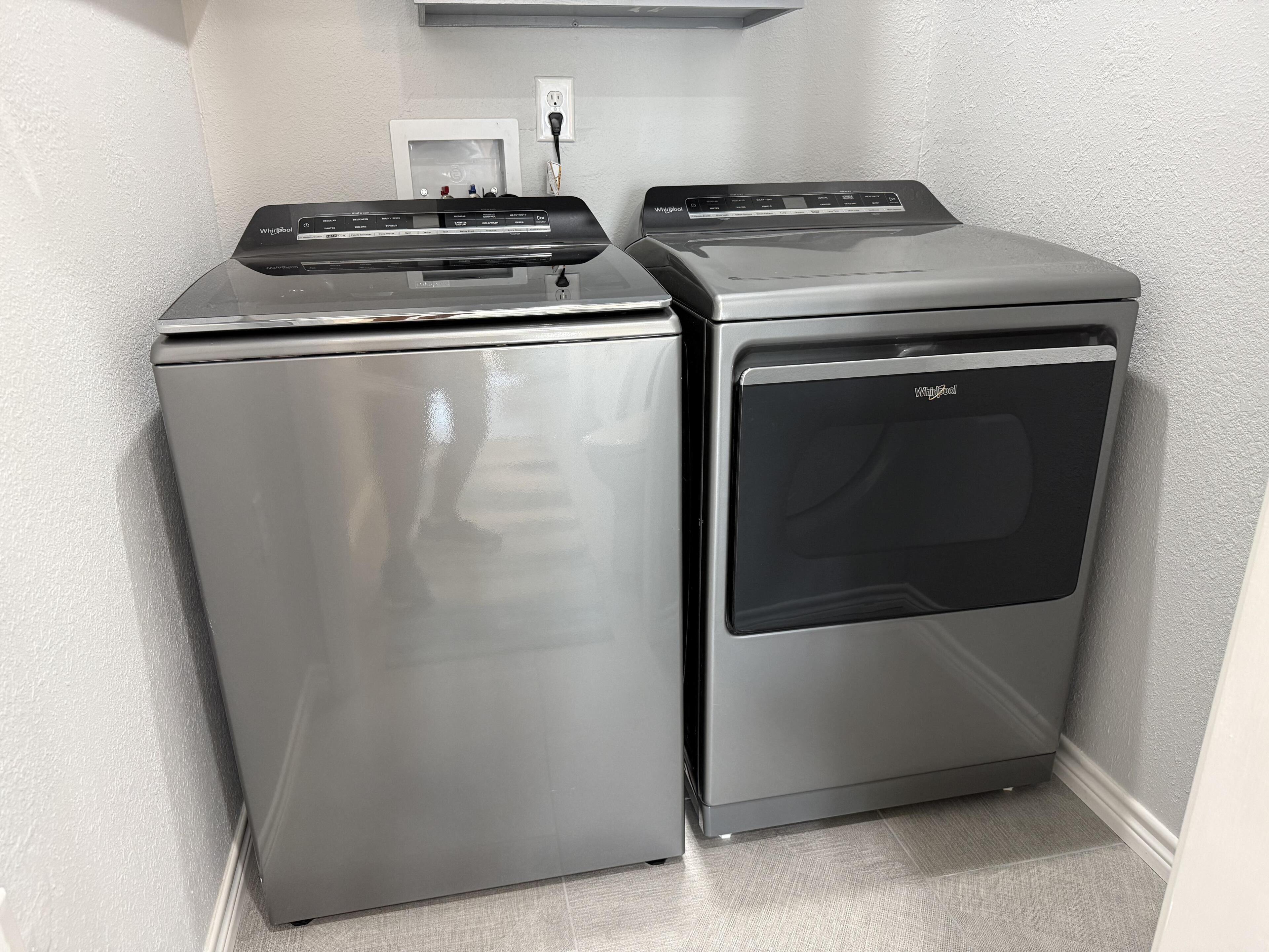 Zwei Edelstahl-Geräte von Whirlpool, eine Waschmaschine und ein Trockner, stehen in einer Ecke mit einer weißen, strukturierten Wand und einer Steckdose darüber.
