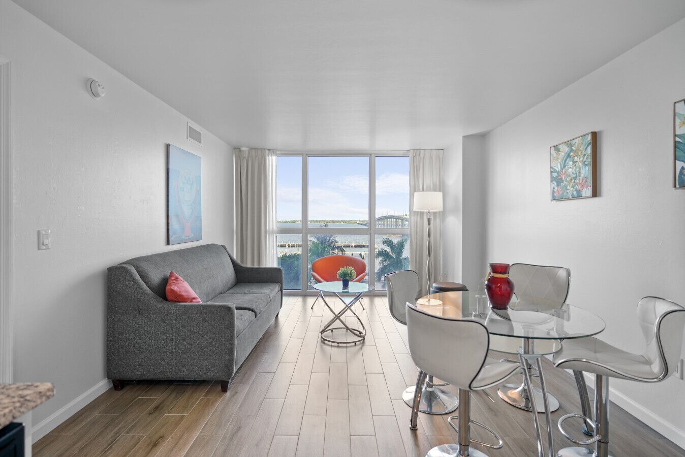 Una sala de estar moderna con un sofá gris, una mesa de comedor de vidrio con sillas y vistas al exterior a través de amplios ventanales.