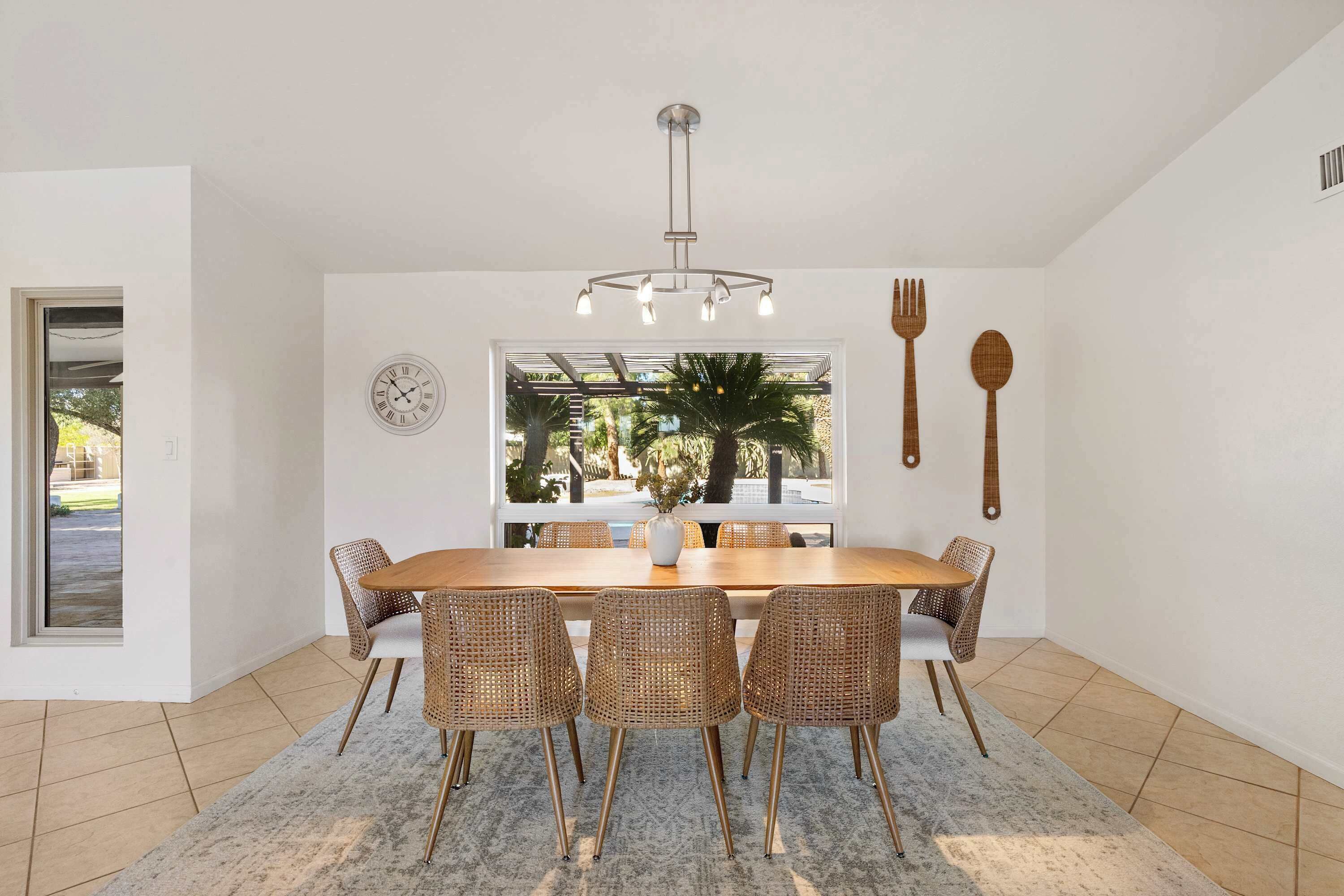 Ein Essbereich mit Holztisch, Korbstühlen, einer modernen Chandelier und einem großen Fenster mit Blick auf Palmen.
