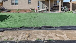 Een gelijkvloers huis met een overdekt patio, een kleine voortuin met kunstgras en een zichtbare airconditioning op het dak.