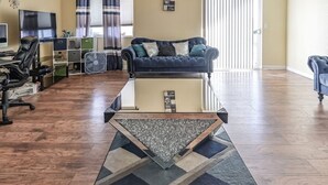 Een ruime woonkamer met een plafondventilator, een groot raam met gordijnen, een open haard en een eethoek met een tafel en stoelen.