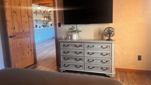 Quarto com porta de madeira, cômoda com várias gavetas, televisão de tela plana fixada na parede e um ventilador sobre a cômoda.