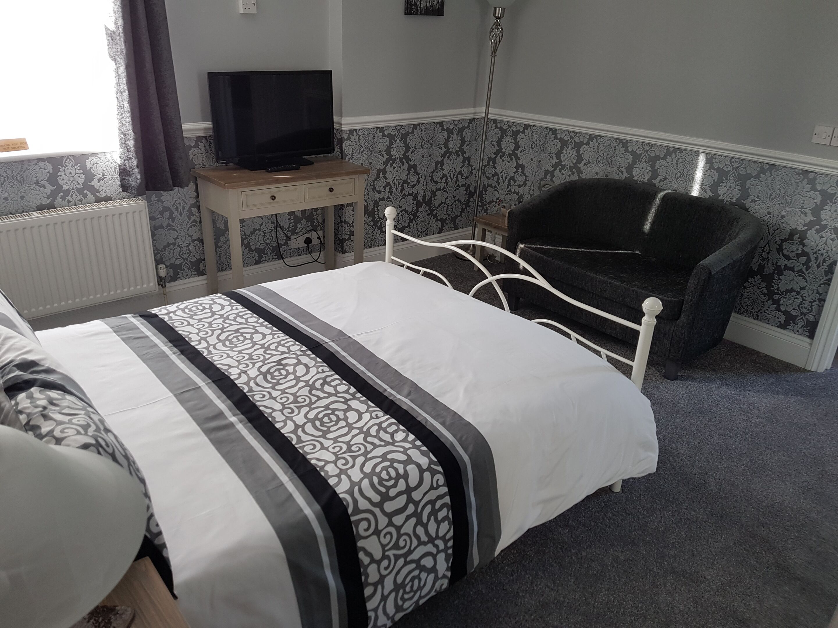 A bedroom with a bed, a chair, a television on a stand, and patterned wallpaper.
