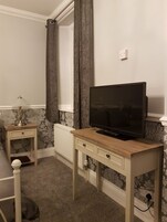 Una habitación con una televisión sobre un mueble de madera, una mesita de noche con una lámpara, un radiador y papel tapiz estampado.