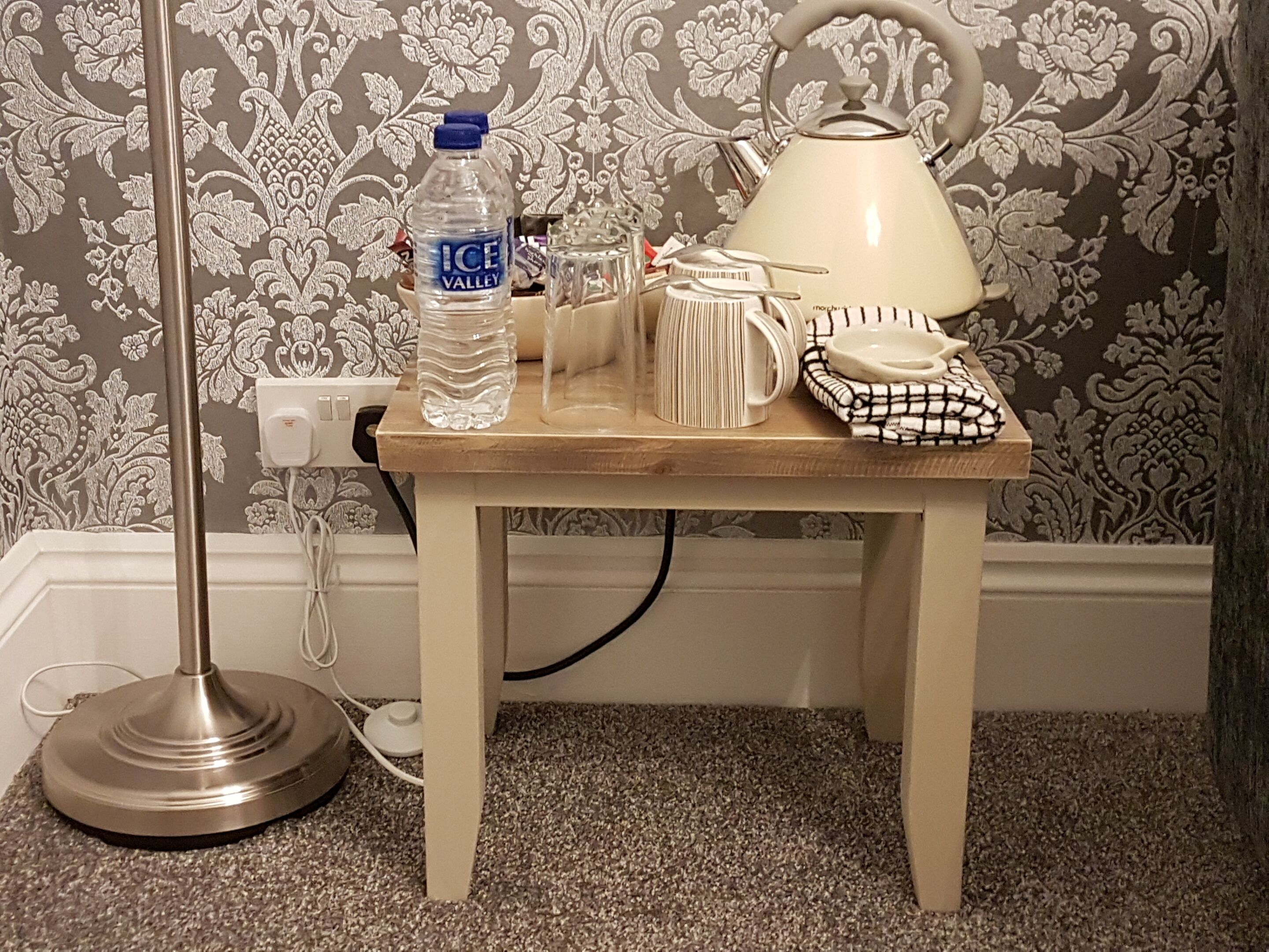 Una pequeña mesa de madera con una lámpara, una botella de agua, una taza y un mantel a cuadros.