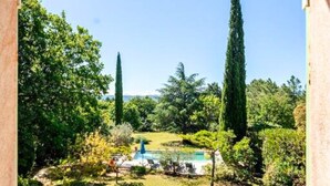 Blick aus einem Fenster auf einen Garten mit Schwimmbad, hohe Bäume und einem klaren Himmel.