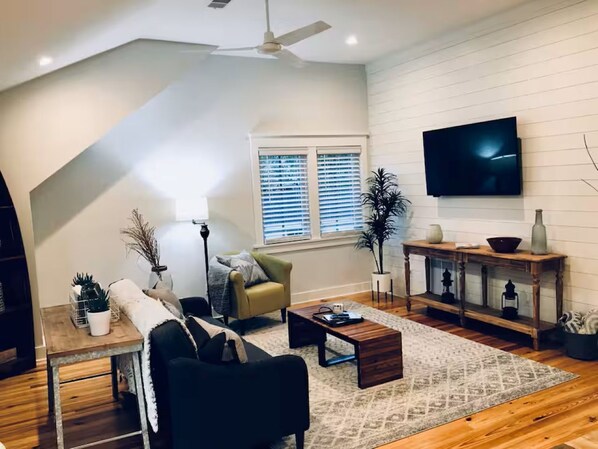 Ein modernes Wohnzimmer mit einer Couch, einem Couchtisch und einem Fernseher. Es gibt ein Fenster mit Jalousien, einen Deckenventilator und einen Holztisch mit Dekoration.