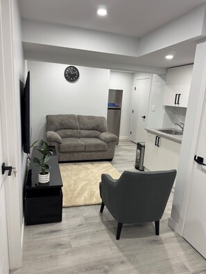 A modern living room with a grey sofa, a black TV stand, and a grey armchair. There is a kitchen area with white cabinets and a stainless steel refrigerator.