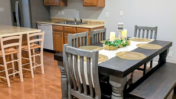 Un comedor con una mesa de madera, sillas y un banco. Se ve una cocina con armarios de madera y electrodomésticos al fondo.