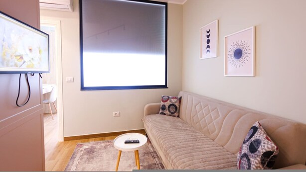 Ein modernes Wohnzimmer mit einem beigefarbenen Sofa, einem kleinen weißen Tisch und einer Fernbedienung darauf. Es gibt ein großes Fenster mit Jalousie sowie zwei gerahmte Kunstwerke an der Wand.