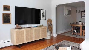 A living room with a flat-screen TV, a wooden cabinet, a dining table with chairs, and a chandelier. - Downtown Ely Home • BWCA Gateway • Walk to Shops (Ely)