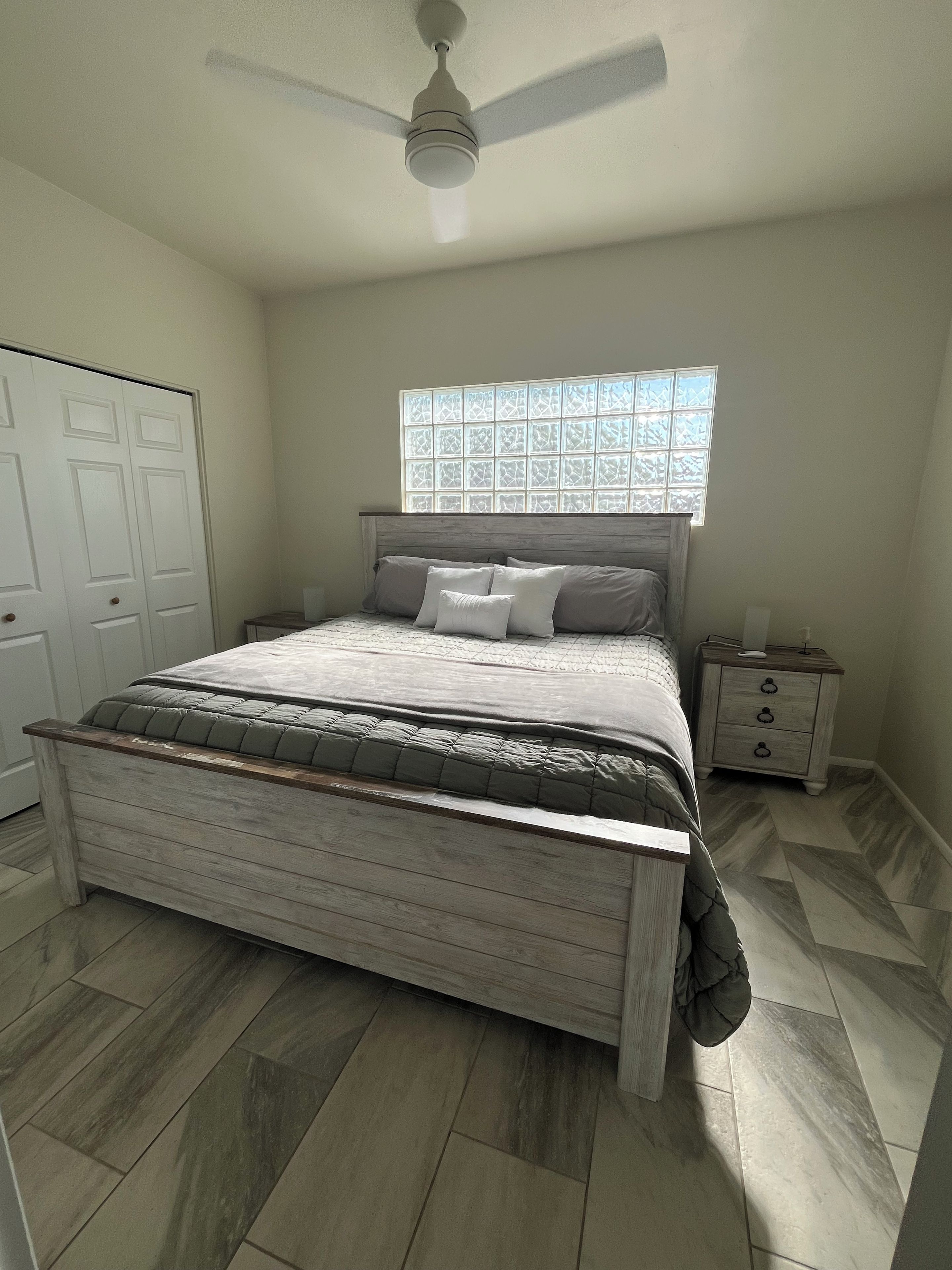 Un dormitorio con cama, mesita de noche, ventana y ventilador de techo.