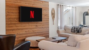 A modern living room with a wooden wall, a mounted TV displaying the Netflix logo, a sofa, a coffee table, and a chair. - Jamaica Chalet: 3 Kings + Arcade | Starlink (Jamaica)