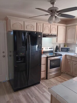 A kitchen with light wooden cabinets, a black refrigerator, a microwave, an oven, and a ceiling fan.