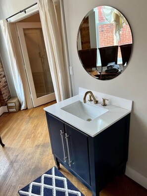 A bathroom with a white sink, dark cabinets, and a round mirror on the wall. - Charming Loft-Style Condo in Beautiful Historic Building (Greenville)