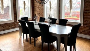 Un coin repas avec un mur en briques, de grandes fenêtres ornées de rideaux blancs, une table en bois et des chaises noires.