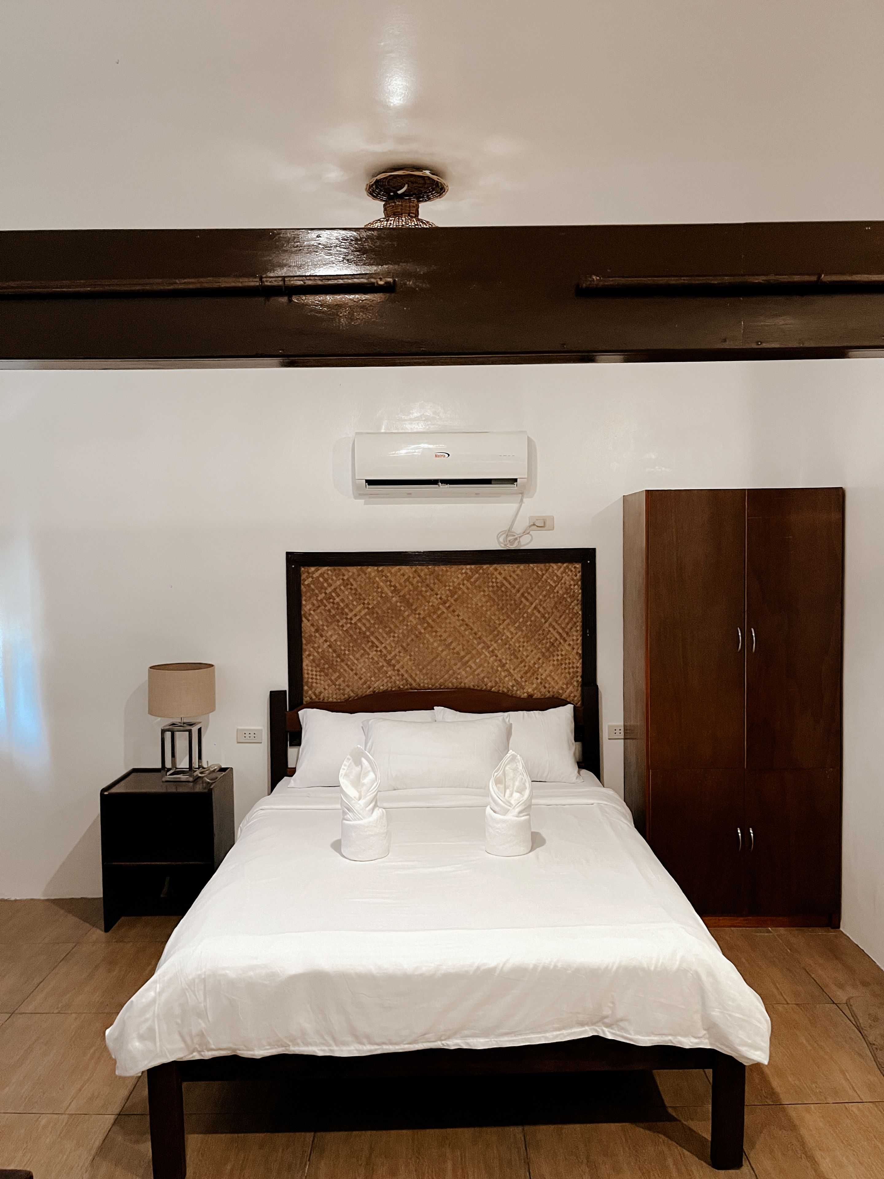 A hotel room with a bed, bedside table, lamp, air conditioning unit, wardrobe, and a ceiling light fixture.