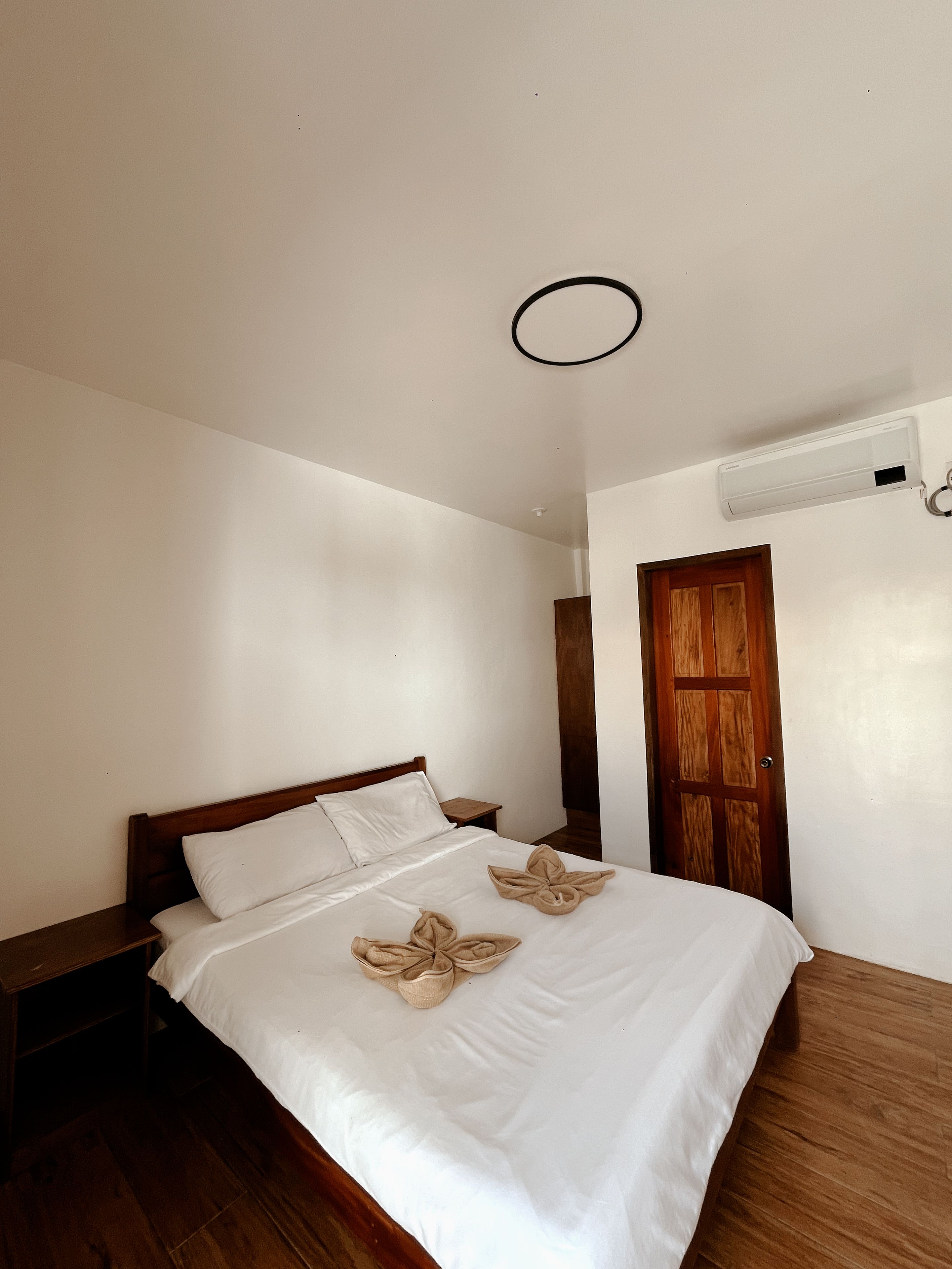 Une chambre d’hôtel avec un lit en bois, du linge de lit blanc, une table de chevet en bois, une porte en bois et un luminaire au plafond.