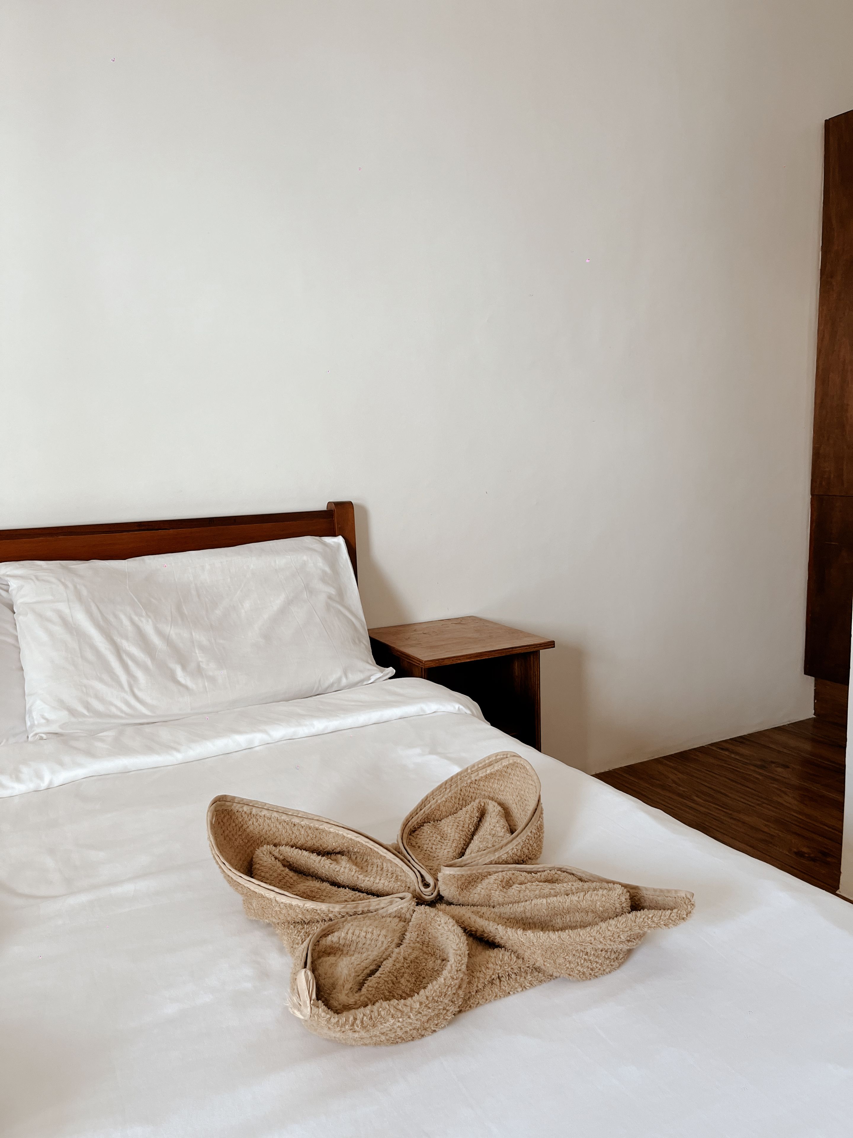A bed with a wooden headboard, a white pillow, and a beige towel arranged in a bow shape.
