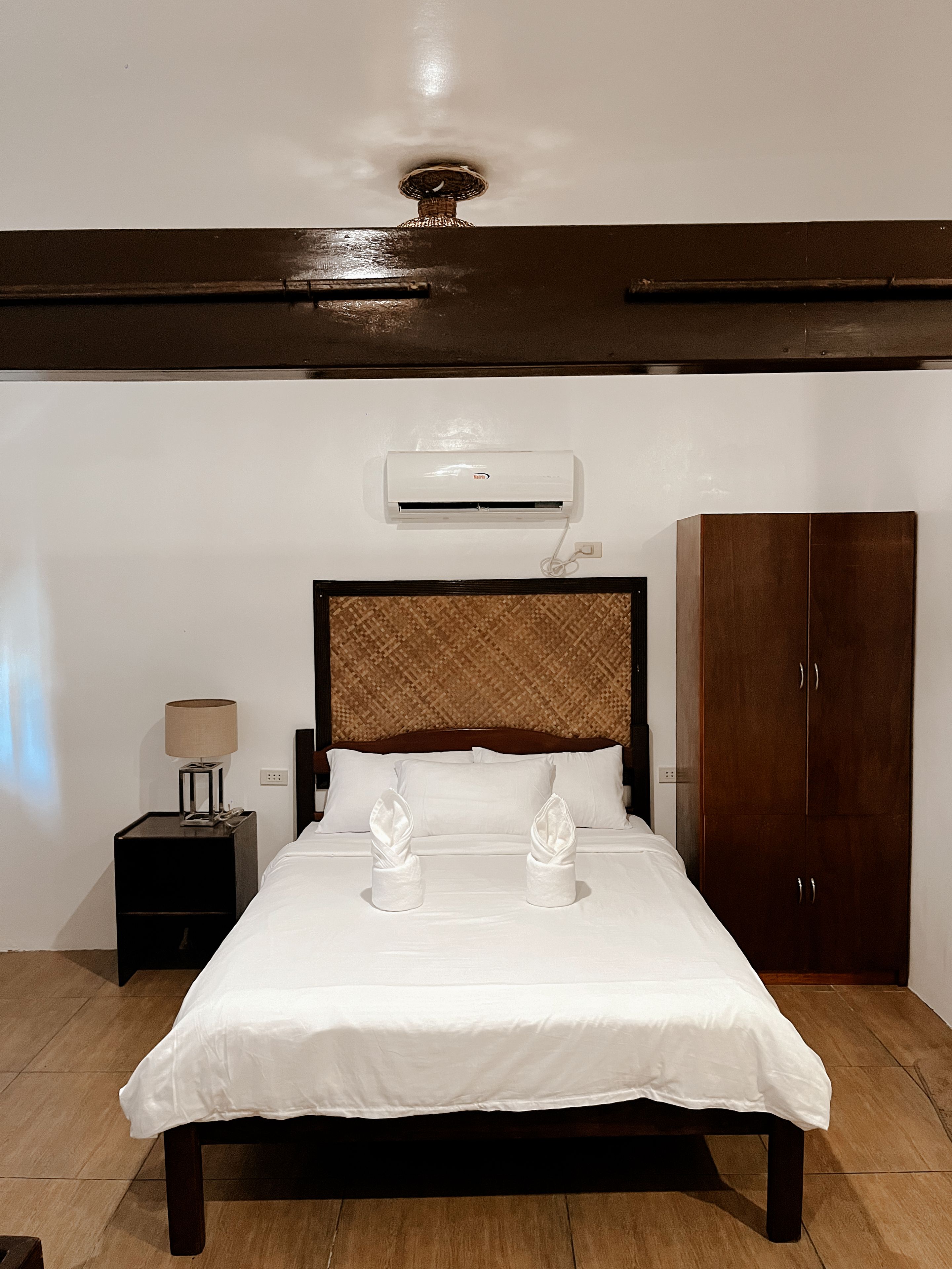 Quarto de hotel com cama, mesa de cabeceira, abajur, guarda-roupa e aparelho de ar condicionado.