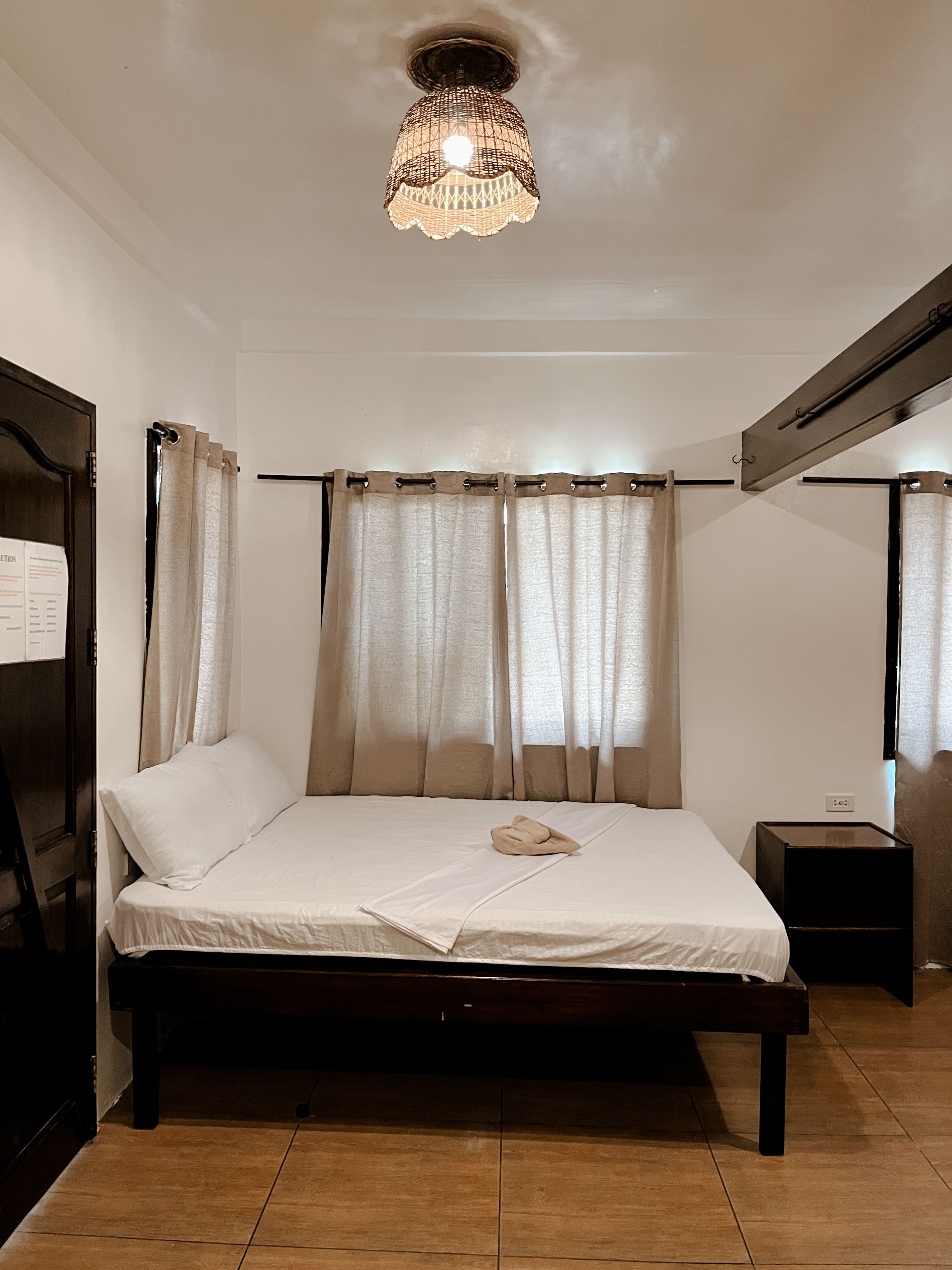 Une chambre à coucher comprenant un lit, des rideaux, une table de chevet, un radiateur fixé au mur et un luminaire au plafond.