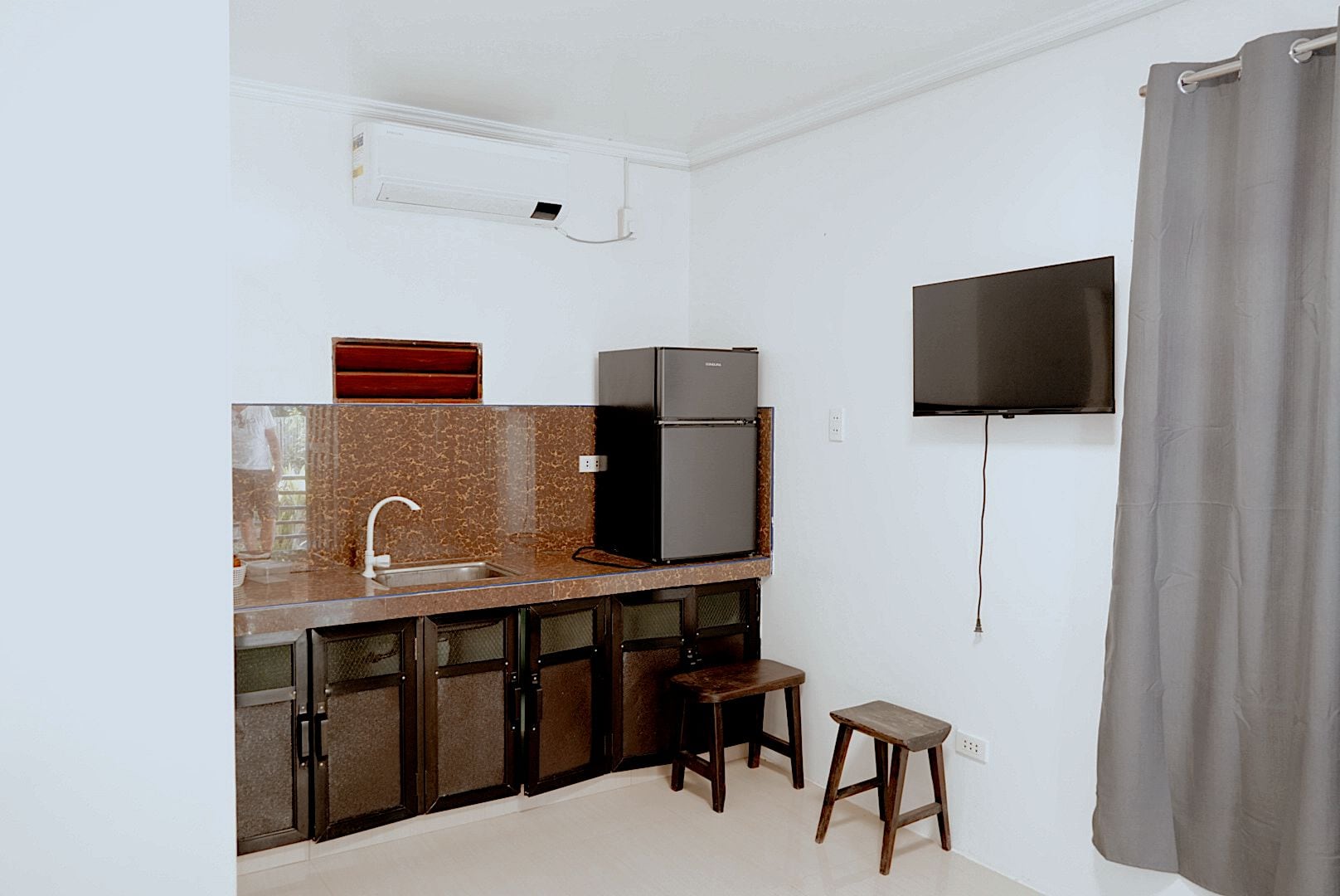 A compact kitchen with a built-in refrigerator, a microwave, and a sink. There is a small stool and a television mounted on the wall.