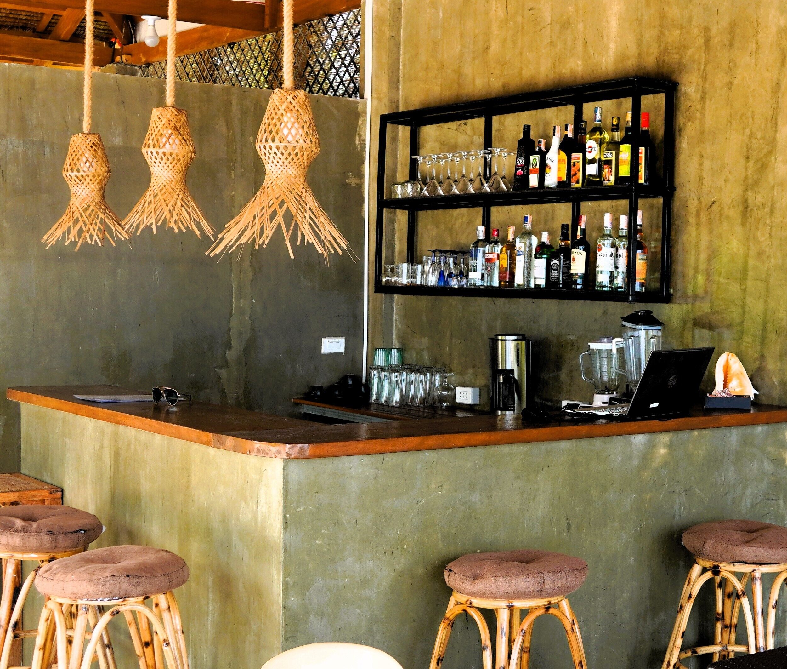 A bar area with stools, a bar counter, hanging lights, and a shelf with bottles and glasses.