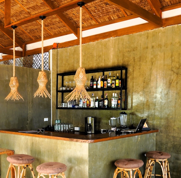 A bar area with hanging rattan light fixtures, a shelf with liquor bottles, a coffee machine, and stools.