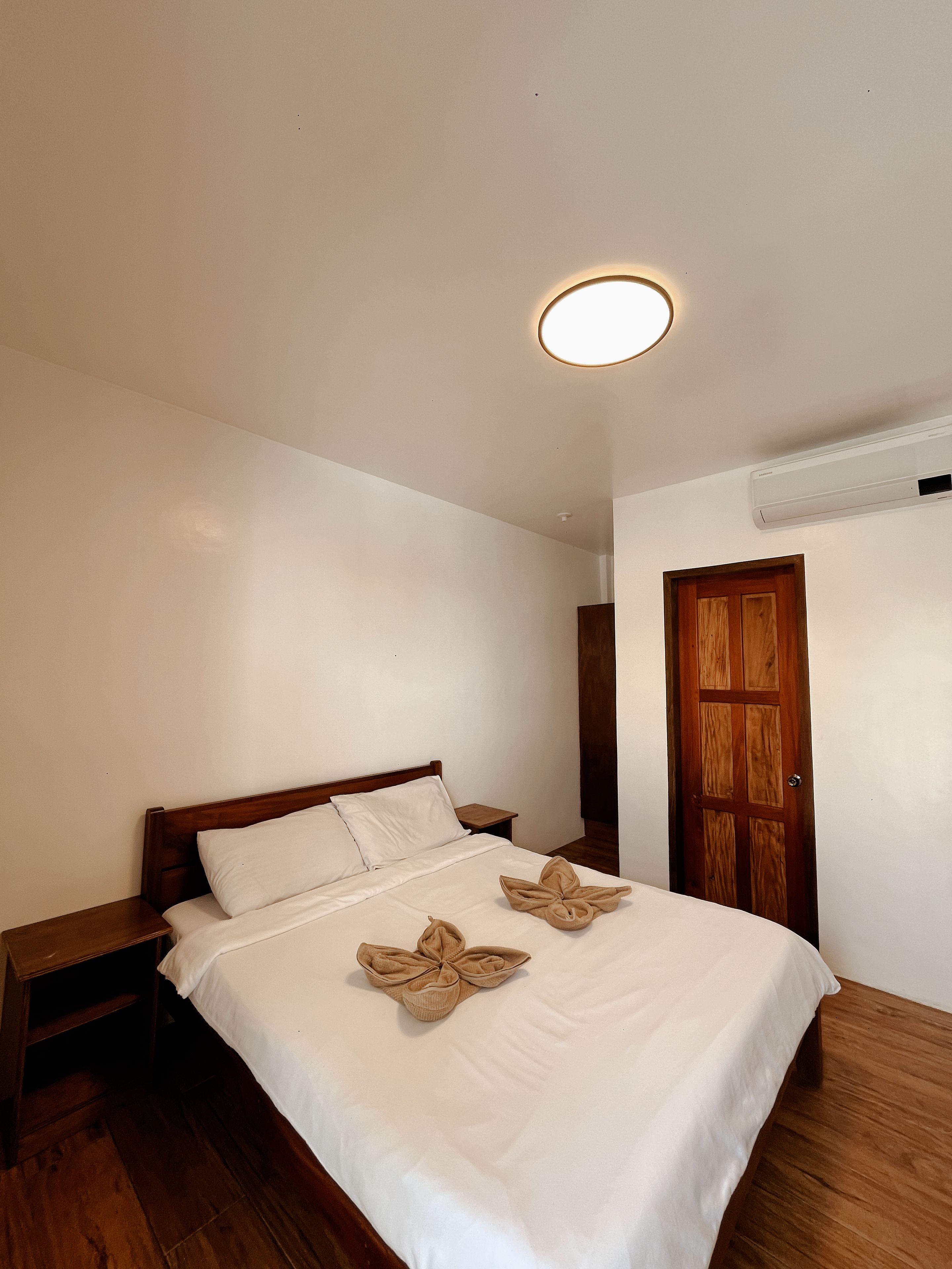 Een slaapkamer met een houten bed, een nachtkastje, een houten deur en een plafondlamp.