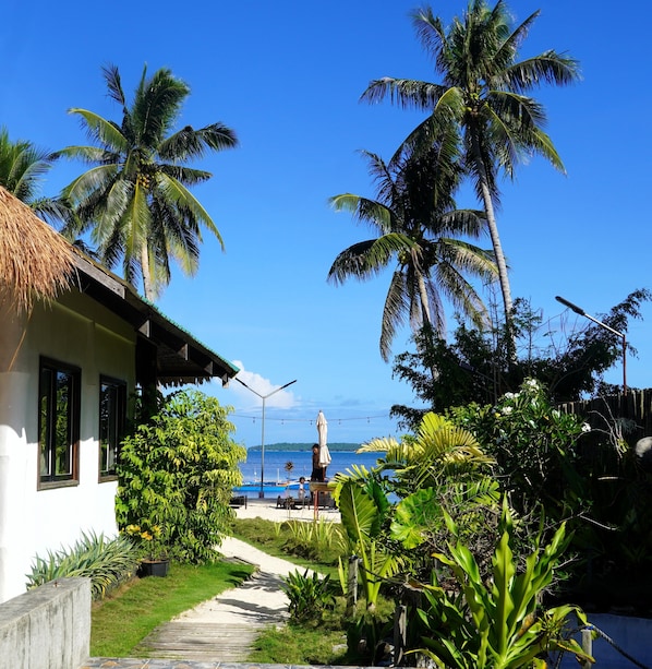 Un resort tropical con una cabaña de techo de paja, palmeras, un camino que lleva a la playa y vista al océano.