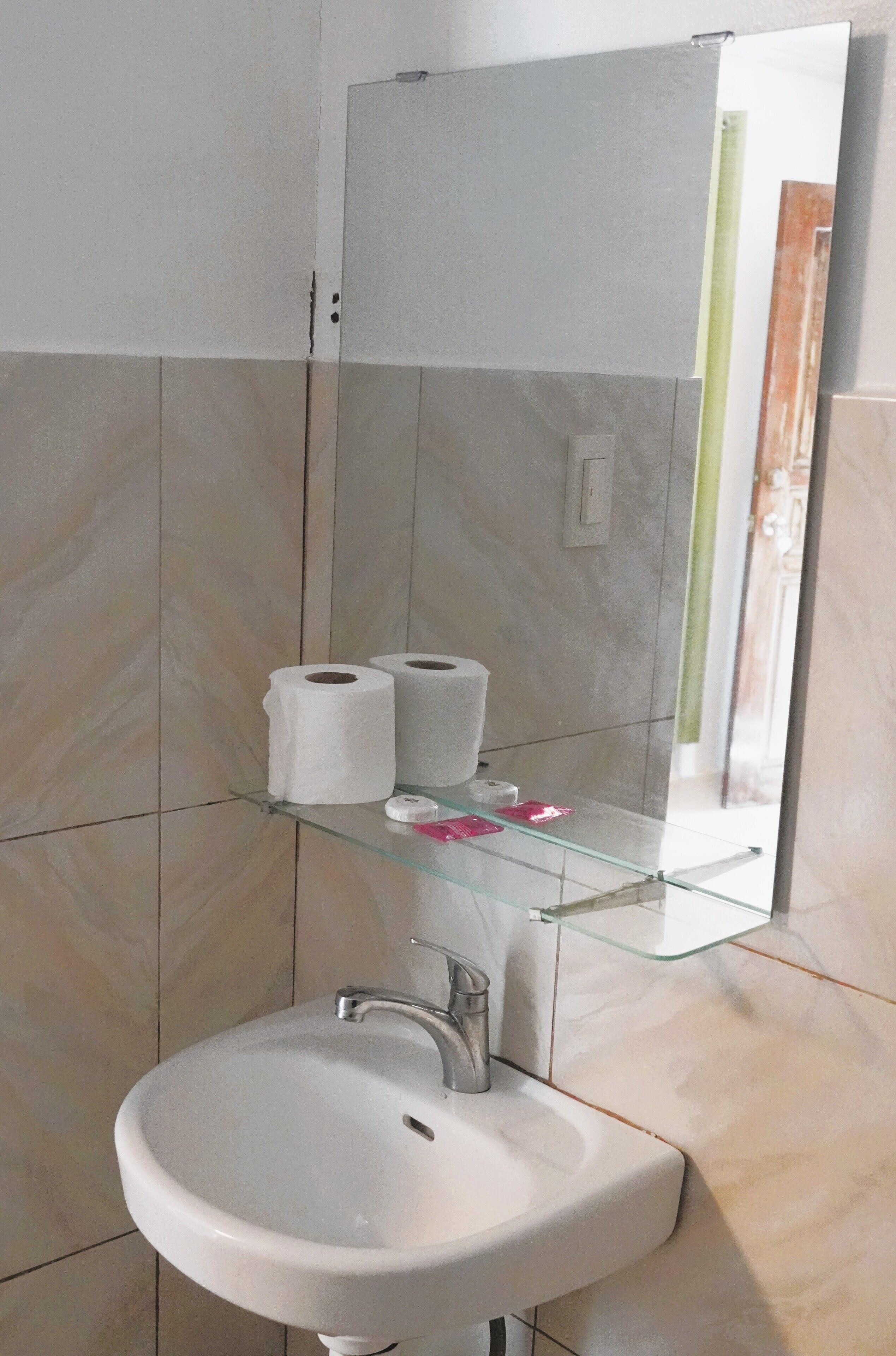 Un baño con un lavamanos blanco, dos rollos de papel higiénico y un estante de vidrio con un cepillo de dientes y un objeto rosa.