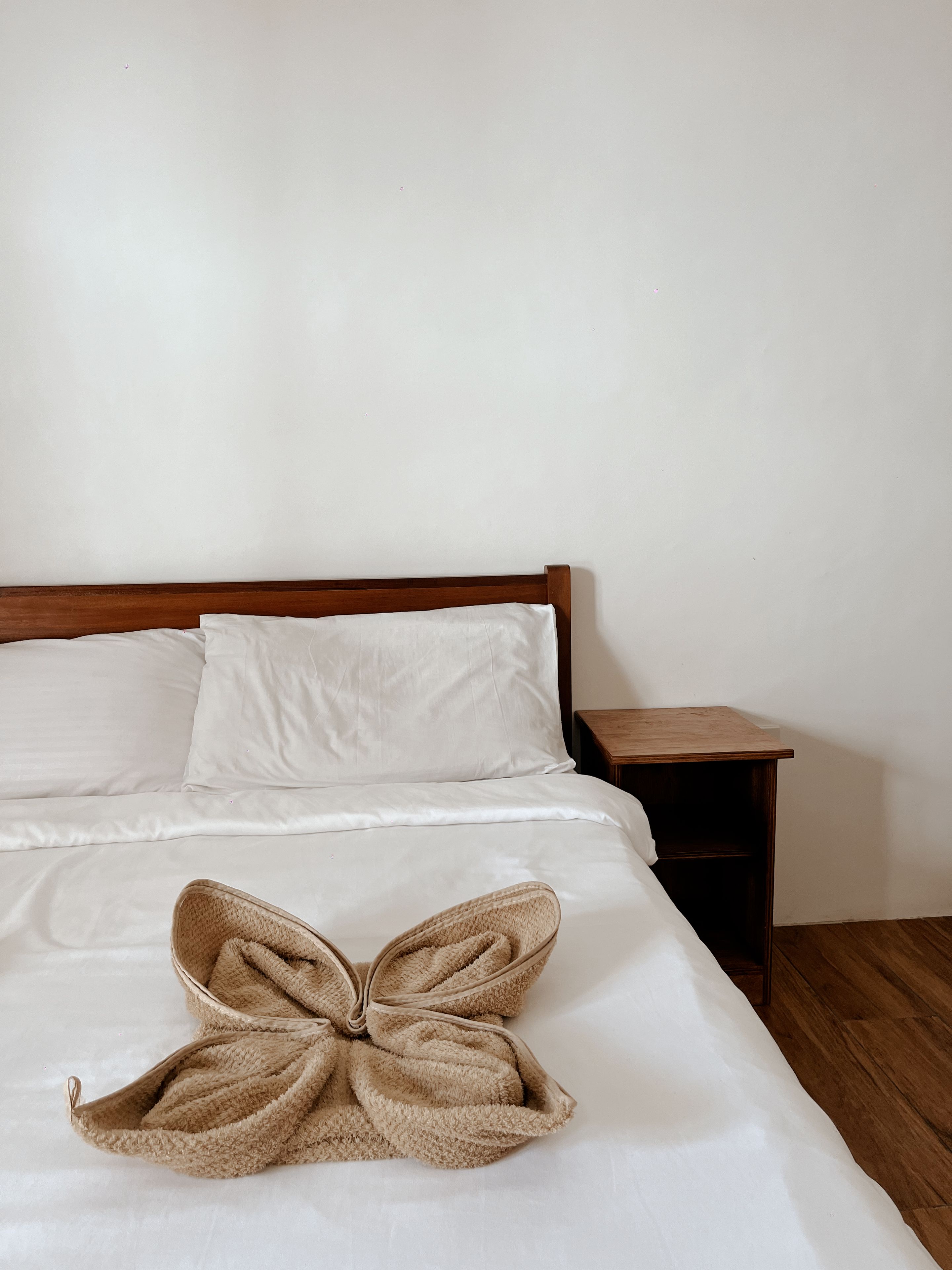 A bed with white linens, a wooden headboard, a wooden nightstand, and a beige towel arranged in a bow shape.