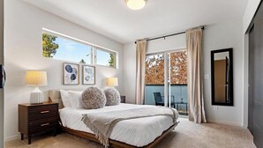Ein Schlafzimmer mit einem großen Bett, zwei Nachttischlampen, einem Nachttisch und einem Fenster mit Blick auf Bäume.