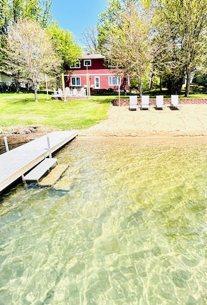 A clear swimming pool with a wooden deck, a red house in the background, and a grassy area with white chairs. - Sunset Beach Getaway - Game Room+XBox+Fire Pit+Fun (Detroit Lakes)