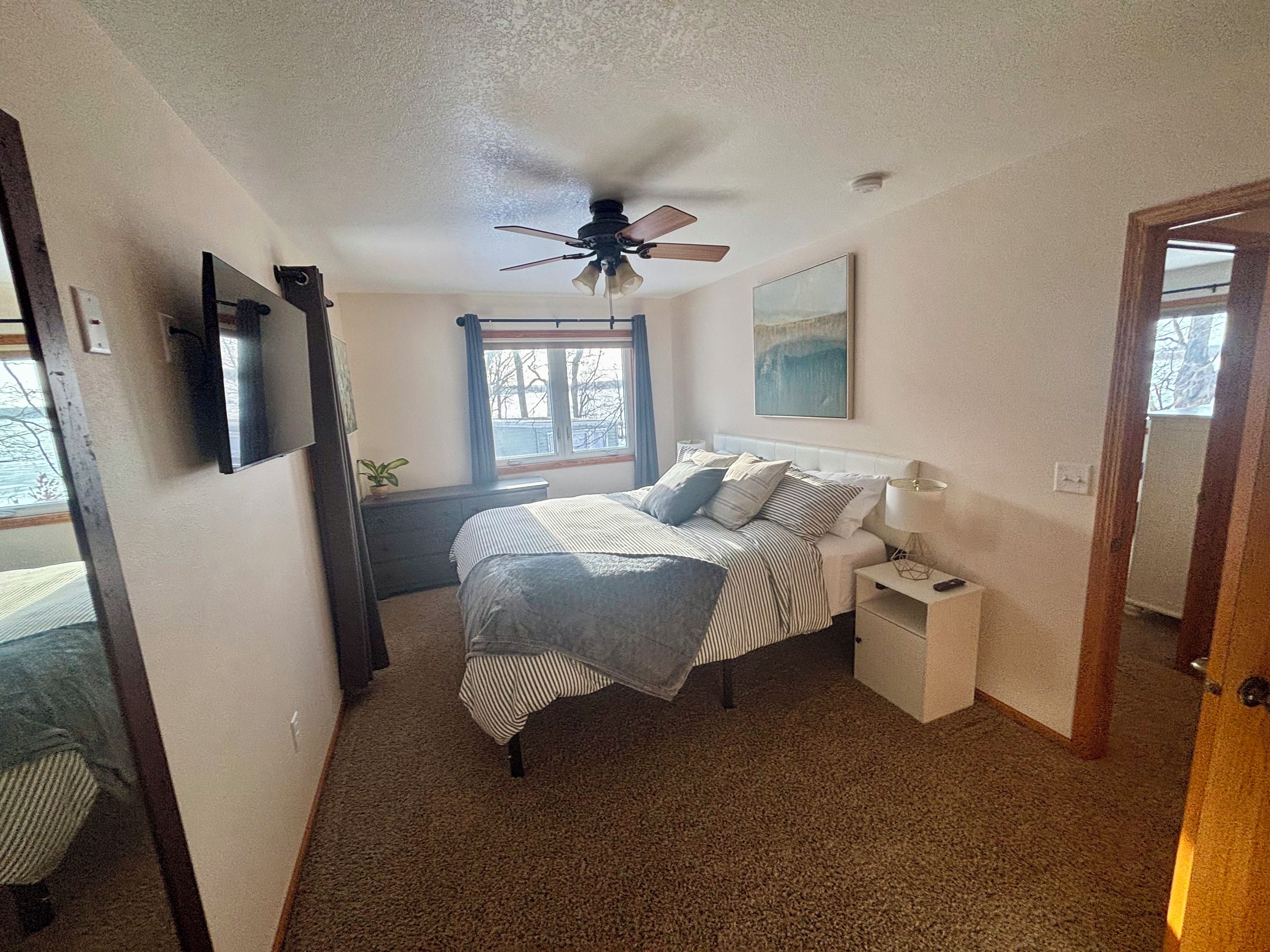 Une chambre à coucher comprenant un lit, une table de chevet, une lampe, un ventilateur de plafond, une fenêtre et un tableau accroché au mur.