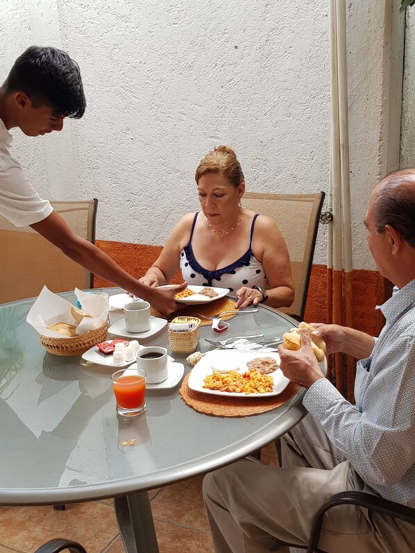 A round glass table with breakfast items, including bread, eggs, and juice, set against a white textured wall. - HOTEL QUINTA REAL LAS PALMAS MALINALCO (Malinalco)