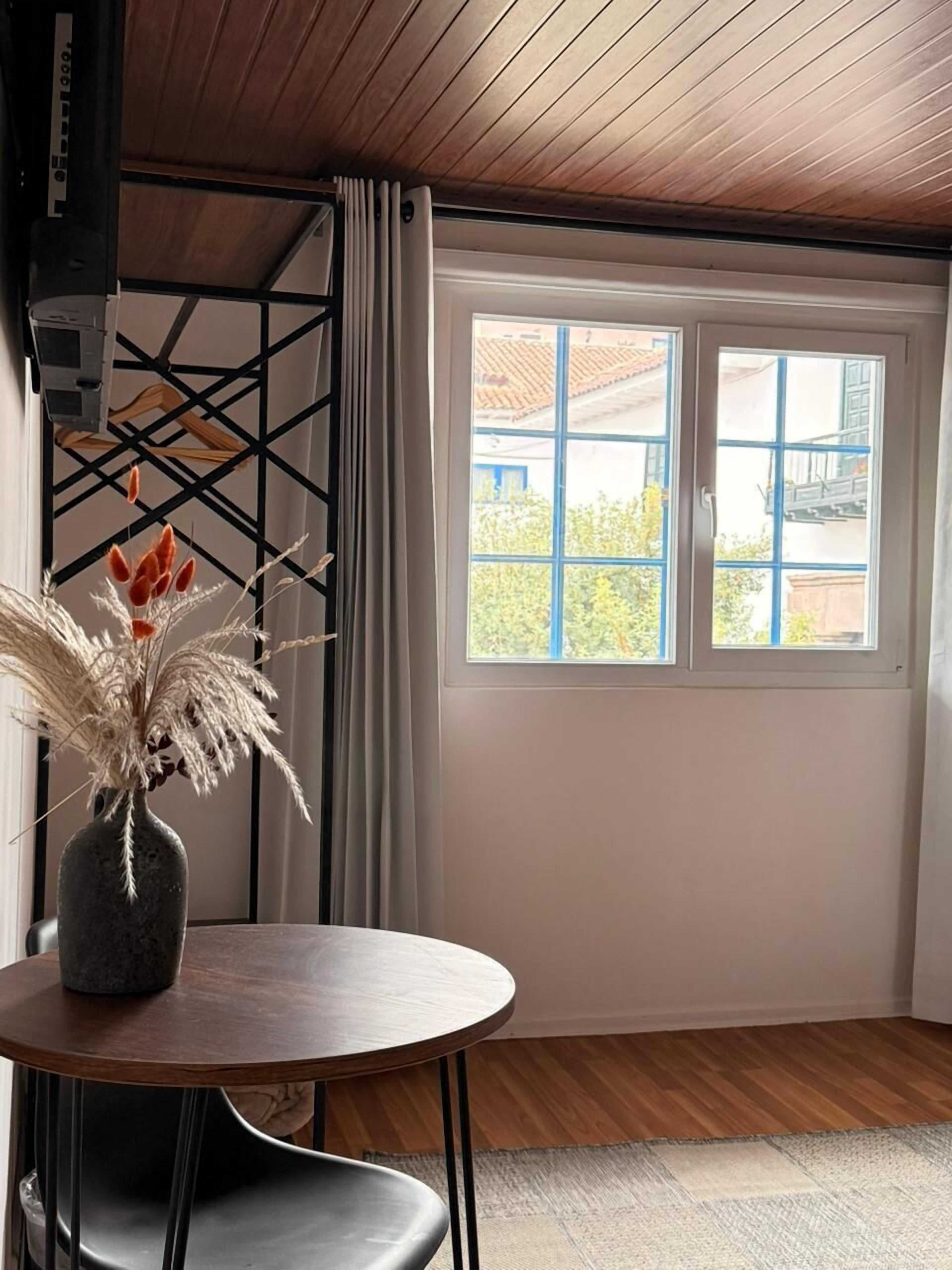 Quarto com teto de madeira, janela com persianas azuis, mesa redonda com um vaso de plantas secas e uma cadeira preta.